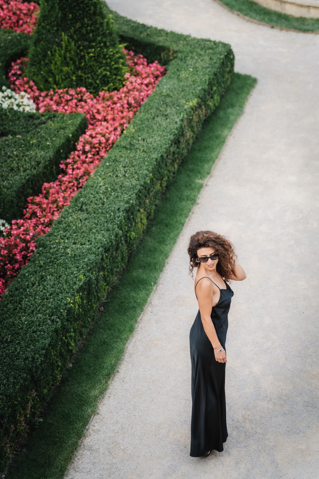 Woman in a long black dress walking a garden path beside pink flower beds in Prague