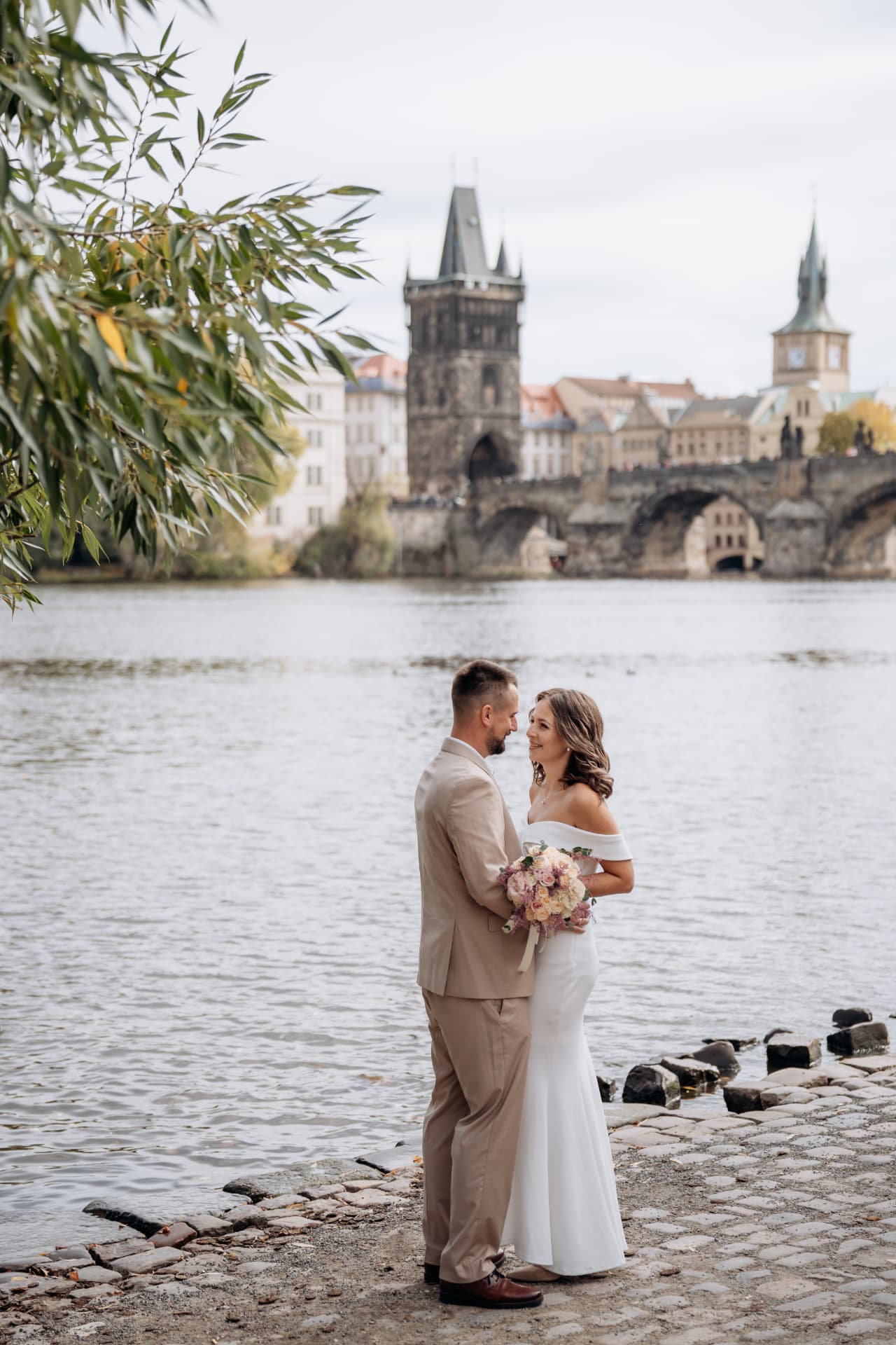 Bride and groom looking into each other's eyes on a cobblestone riverbank beneath the arches of Charles Bridge in Prague