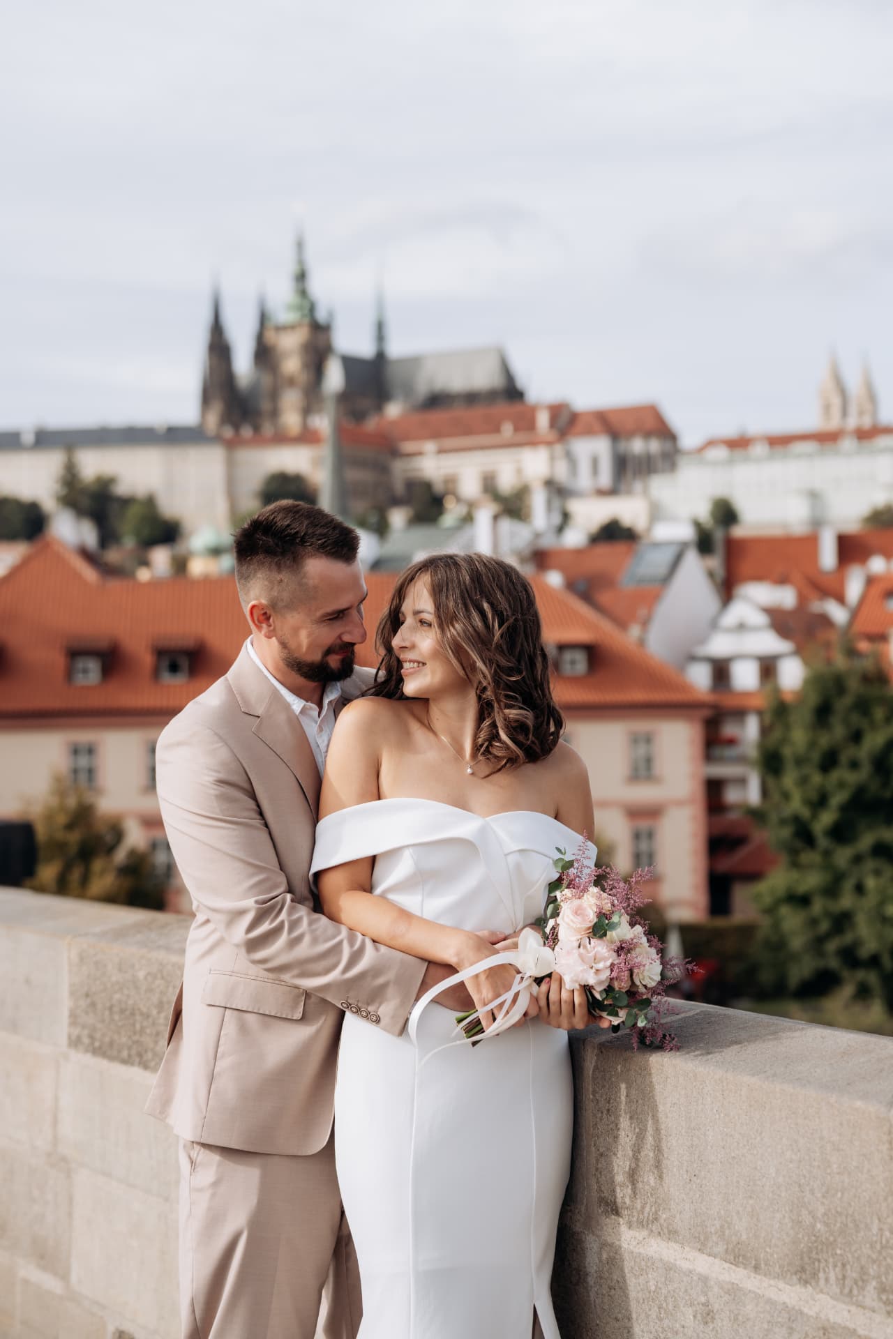 Wedding photoshoot in Prague of a bride and groom embracing at a stone parapet with Prague Castle and St. Vitus Cathedral behind them