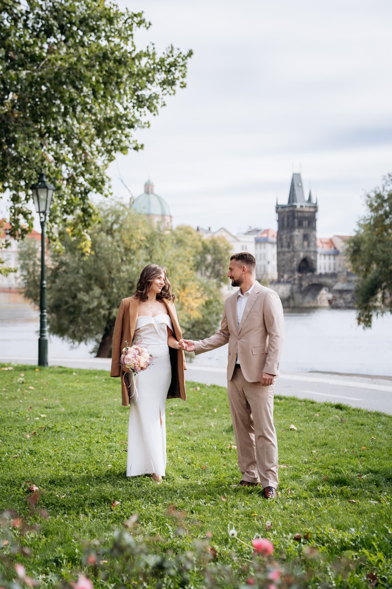 Wedding couple holding hands on a riverside lawn in Prague with the Old Town Bridge Tower behind them