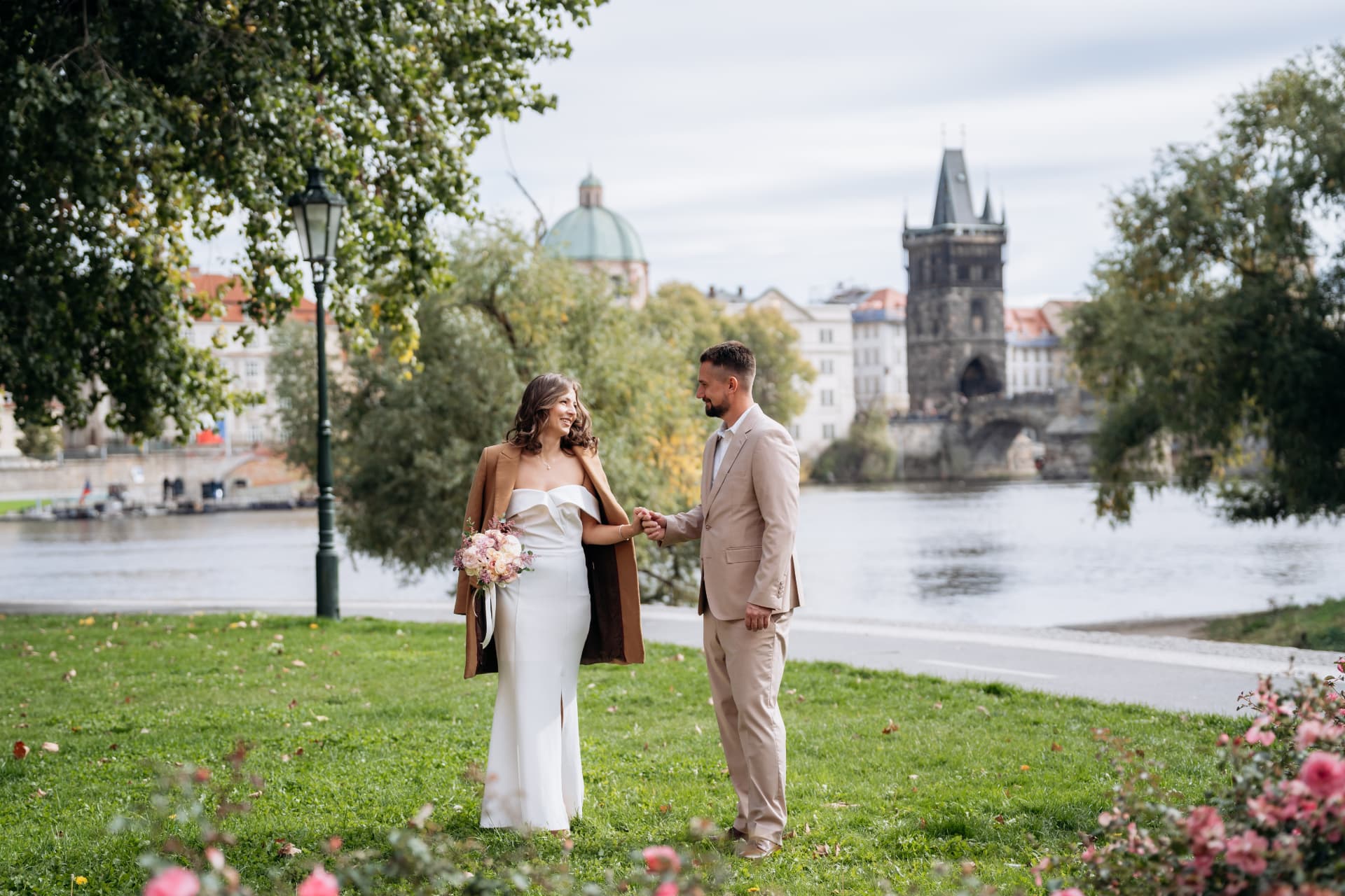 Bride and groom holding hands on a lawn by the Vltava river with Charles Bridge and the Old Town Bridge Tower in the background